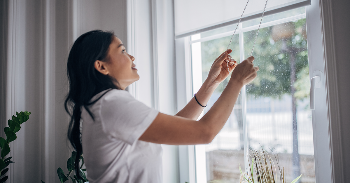 A smiling person adjusts the window blinds so daylight can pour into a home.